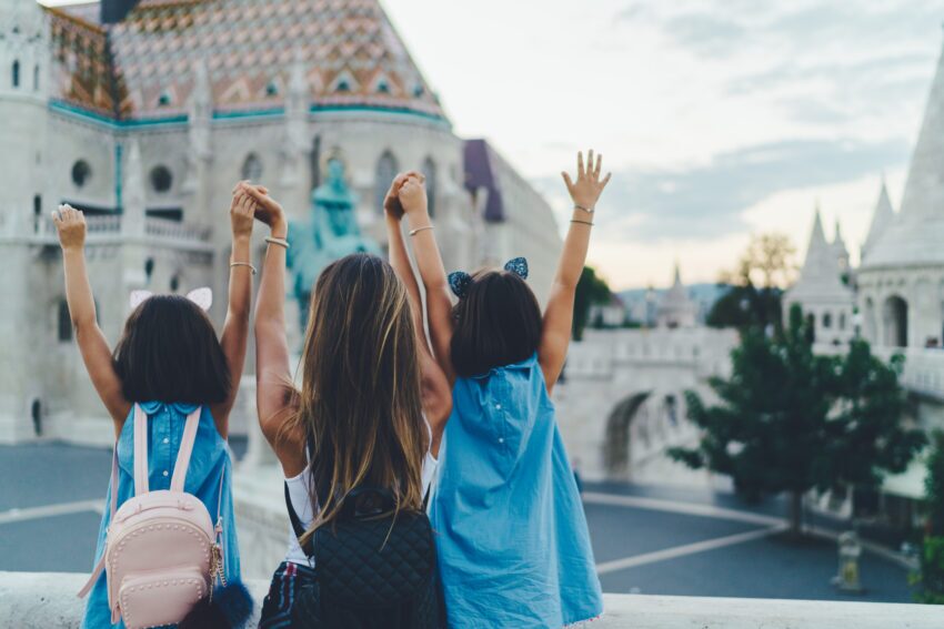 A mother and two daughters viewed from behind with their arms raised while viewing a Budapest city street