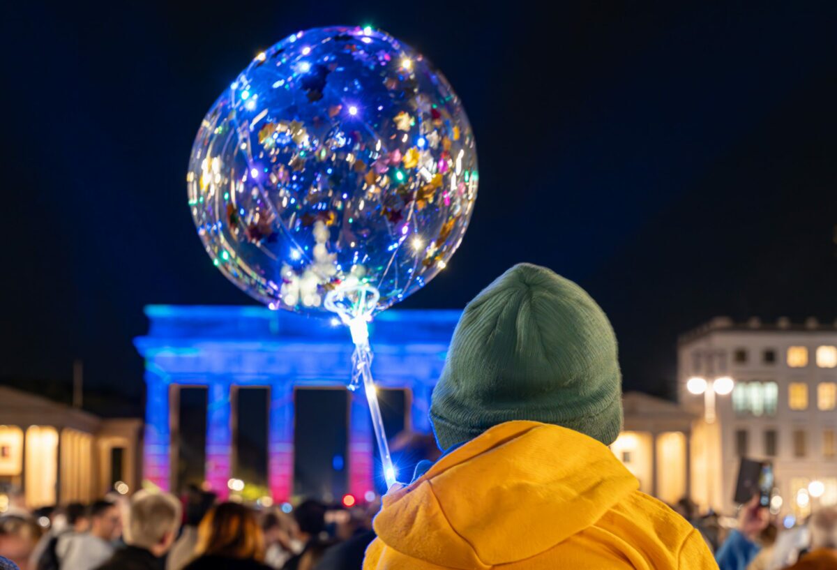 Rear view of a kid with yellow jacket and green hat holding a lit-up balloon against Berlin Brandenburg Gate at Festival of Lights