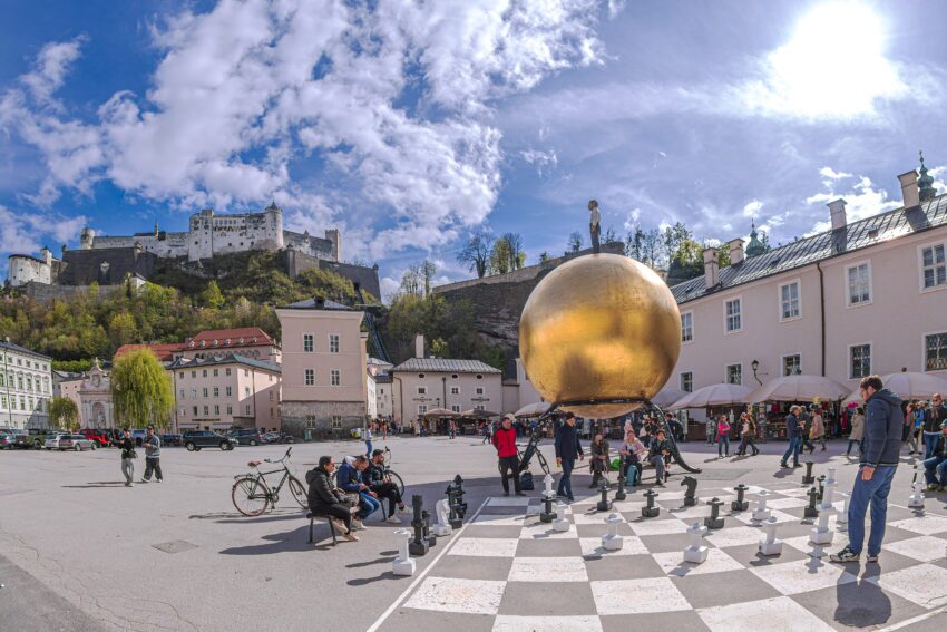 Salzburg: Chess game drawn on asphalt in Kapitelplatz, near monument to Paul Fuerst "Sphere", a golden ball on which stands a small man. Castle Hohensalzburg in background.
